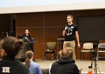Kurt Lewis, computer science class of 2019, is speaking with a microphone in front of a crowd of students in an auditorium before a hackathon.