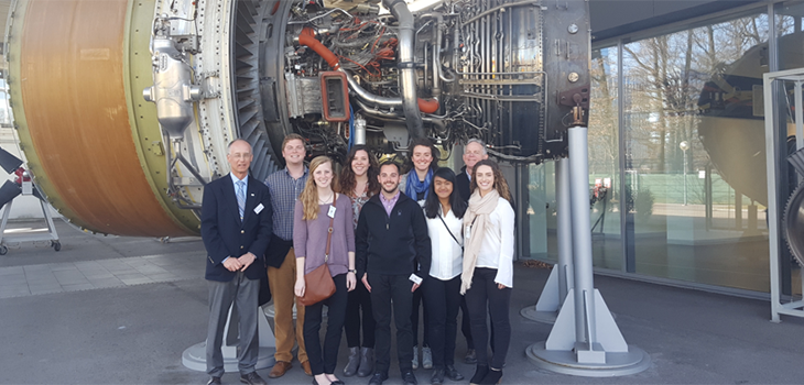 Eight UC students pose with a guide in front of a jet engine.